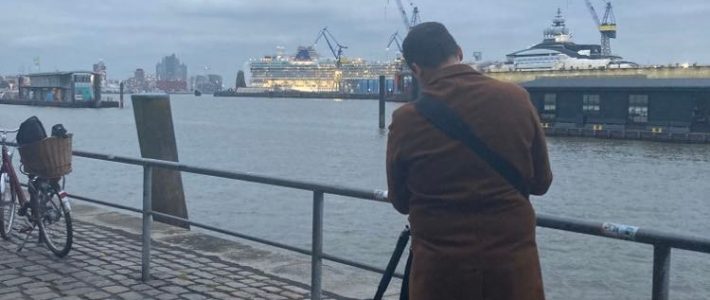 Ein Mensch von hinten mit einem Stativ. Im Hintergrund ist der Hamburger Hafen zu sehen.