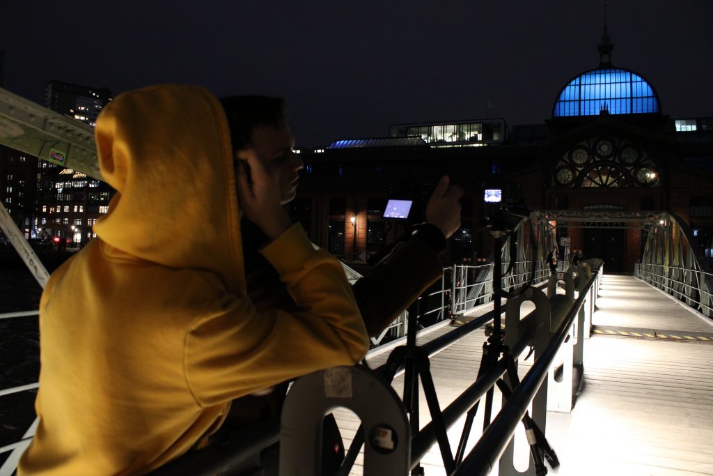Ein Mensch mit gelben Kapuzenpullover fotografiert die Fischauktionshalle bei Nacht. 