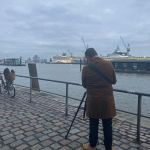 Ein Mensch von hinten mit einem Stativ. Im Hintergrund ist der Hamburger Hafen zu sehen.
